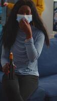 Vertical . African woman keeping social distancing wearing face mask while meeting with friends to prevent spread of coronavirus holding beer bottle looking at camera sitting on couch, covid 19 outbreak video