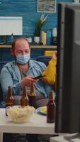 Vertical . Group of multi ethnic friends watching comedy show on tv laughing wearing face mask to prevent infection with covid 19, during global pandemic having fun sitting on couch keeping social distance video