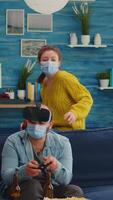 Vertical . Group of multi ethnic friends having fun playing games using virtual reality headset and joystick wearing face mask keeping social distancing. Diverse people enjoying at new normal party video