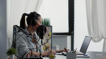 Internet radio show host talking on microphone while checking subject list on laptop. Popular social media content creator speaking to audience while sitting in modern studio. video