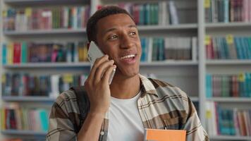 Happy African American man talking mobile phone smartphone technology communicate conversation library student learner university chatting smiling male guy bookshelf educational study calling laughter video