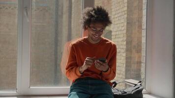 African American student guy using mobile phone relaxing near window in corridor university college school smartphone app chatting smiling happy positive education study break man male use cellphone video