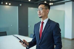 photo of man in navy suit outfit and red tie holding tablet in the office,