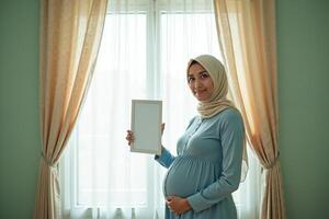 photo of muslim pregnant woman with hijab happy standing holding empty white photograph frame,