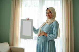 photo of muslim pregnant woman with hijab happy standing holding empty white photograph frame,