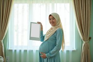 photo of muslim pregnant woman with hijab happy standing holding empty white photograph frame,