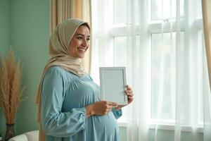 photo of muslim pregnant woman with hijab happy standing holding empty white photograph frame,