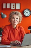 A smiling middle aged woman in an orange office, seated at her desk with a laptop photo