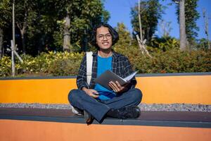A male student is sitting in the park and holding a book while smiling. photo