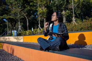 Male student is sitting in the park and holding a book with a thoughtful expression. photo