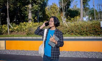 A male student wearing a plaid shirt and glasses is standing in the park answering a phone call and holding a book. photo