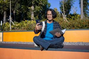 A male student wearing a checked shirt and glasses sits in the park taking a selfie with his phone. photo
