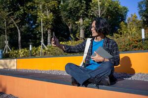 A male student wearing a checked shirt and glasses sits in the park taking a selfie with his phone. photo