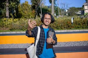 A male student wearing a shirt and glasses in the park is holding a book while making an inviting gesture. photo