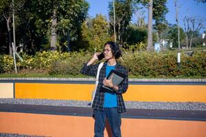 A male student wearing a plaid shirt and glasses is standing in the park answering the phone while smiling. photo