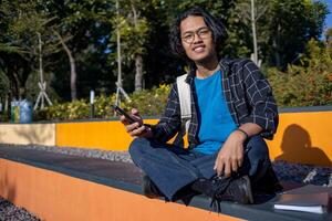 A man wearing a plaid shirt sits in the park while playing with his cellphone with various expressions. photo