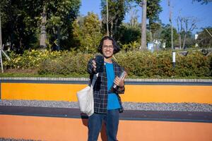 A male student wearing a plaid shirt and glasses is standing in the park showing his cellphone screen. Smiling expression photo