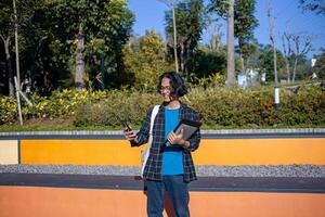 A male student wearing a plaid shirt and glasses is standing in the park smiling while looking at his cellphone screen. photo