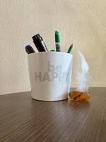 A white mug with the text Be Happy filled with pens and a marker, next to a feather and an amber stone on a wooden surface. photo