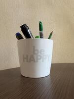 A white ceramic pen holder with Be HAPPY written on it contains various writing instruments including markers and a pencil, sitting on a wooden surface against a textured wall. photo