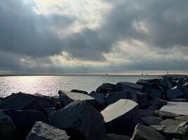 A serene seascape with large rocks in the foreground, distant ships preparing to enter the harbor, under a dramatic cloudy sky with sunbeams shining through. photo
