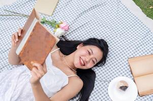 A top view of a positive, lovely Asian woman reading a book and lying on a picnic blanket. photo