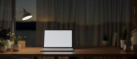 A laptop computer on a wooden table by the window in a house at night, illuminated by a table lamp. photo