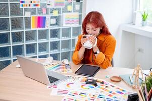 A relaxed Asian female graphic designer is enjoying a morning coffee while working at her desk. photo