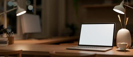 A laptop computer mockup on a wooden table in an office at night, illuminated by table lamps. photo