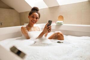 A woman is smiling while bathing in a jacuzzi, using her phone in the bathroom photo