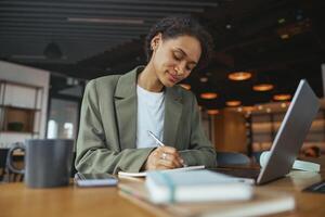 A woman at a desk with a laptop and notebook, working in formal wear in nice modern coworking office photo