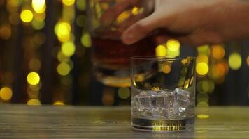 Pouring of golden whiskey, cognac or brandy from bottle into glass with ice cubes. Shiny background video
