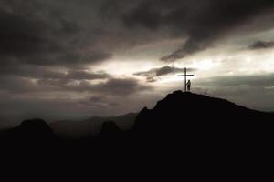 Silhouette of hopeful woman standing alone by cross search for purpose on scenic hilltop viewpoint with dramatic sky background sunshine on sunset.Conceptual loneliness, despair, search for purpose photo