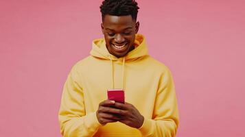 Young man in a yellow hoodie smiles while texting on his pink smartphone against a pink background photo