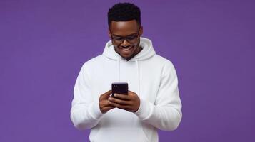 Young man in a white hoodie smiles while using a smartphone against a purple background photo
