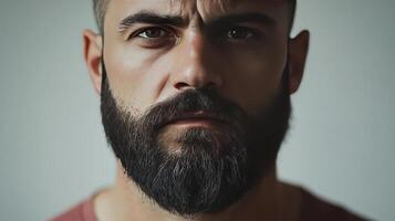 A close-up portrait of a man with a thick beard, neutral expression, and intense gaze, set against a simple background photo
