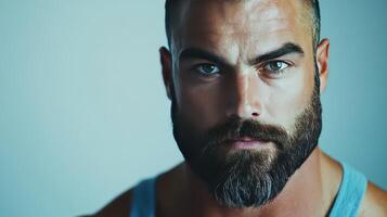 A close-up portrait of a bearded man with striking blue eyes, wearing a tank top, against a simple background photo