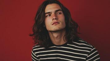 Young man with long hair wearing a striped shirt poses against a red wall in a studio setting during golden hour photo