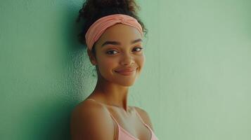 Young woman smiling while leaning against a green wall, wearing a pink headband and top in a bright, cheerful setting photo