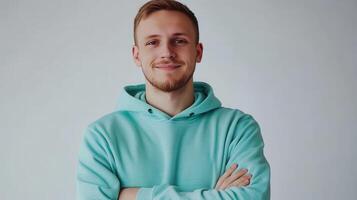 Young man smiling confidently while standing with arms crossed in a light blue hoodie against a plain background photo