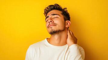 Young man experiencing discomfort while rubbing his neck against a vibrant yellow backdrop in a casual indoor setting photo