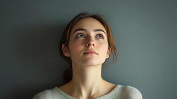 Young woman with brown hair gazes thoughtfully upward against a gray wall in a serene indoor setting photo