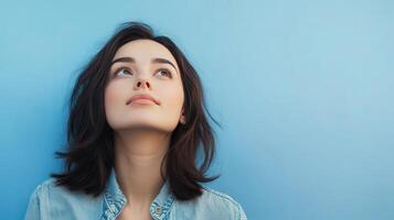 Young woman with dark hair gazing upward against a solid blue background during daylight hours photo