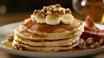 Stack of fluffy pancakes topped with bananas, walnuts, and maple syrup, served with crispy bacon on a plate photo
