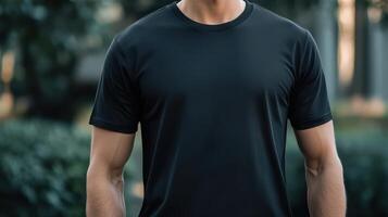 Man in a plain black t-shirt stands outdoors surrounded by greenery in soft sunlight photo