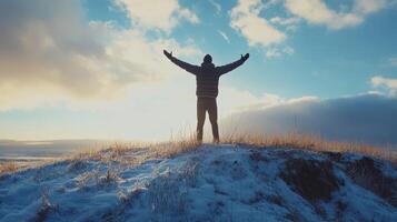 Person standing on a snowy hilltop with arms raised against a bright sky at sunset photo