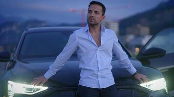 A Stylish Man Is Posing Confidently by a Luxury Car Against the Background of Twilight Sky video