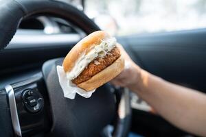 Asian lady holding hamburger to eat in car, dangerous and risk an accident. photo