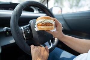 Asian lady holding hamburger to eat in car, dangerous and risk an accident. photo