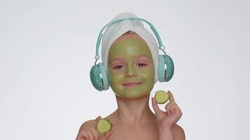 Smiling child girl moisturizing green cucumber mask on face listening to music on headphones dancing video